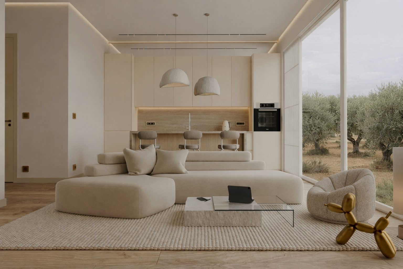 **Alt text (EN):**<br />
Minimalist living room in soft beige tones with a low modular sofa, textured round rug, and a stone coffee table with a glass top. Behind the seating area is an open kitchen with light cabinetry and a natural stone backsplash. Floor-to-ceiling windows look out onto an olive grove. Subtle ceiling lighting and brushed bronze electrical outlets add warm contrast to the light walls.<br />
